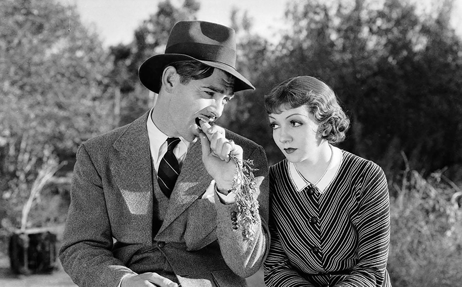 Two people sit outdoors among trees; one wears a suit, tie, and hat while biting a carrot, the other wears a striped dress with a white collar