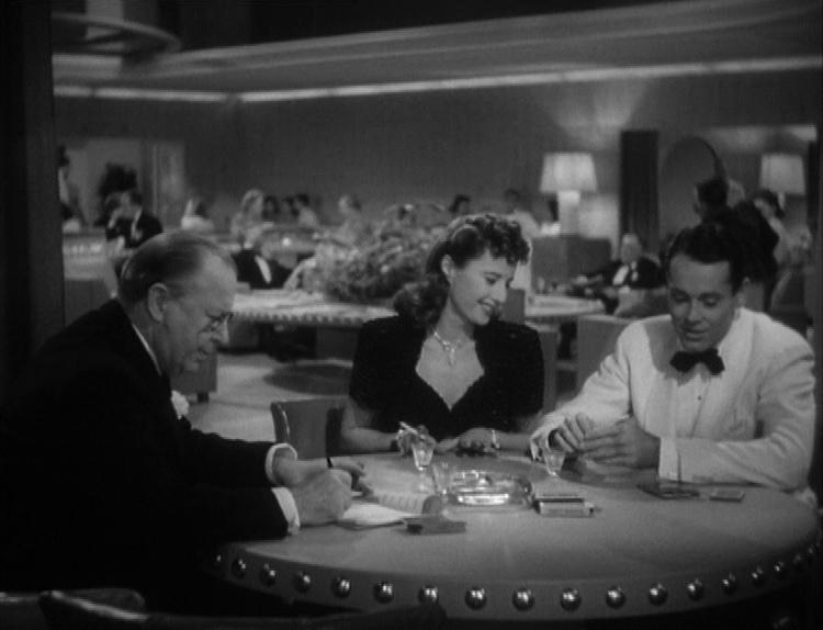 Three people sit at a round table in a casino or upscale club, with drinks and an ashtray; other patrons and ambient lighting are visible in the background