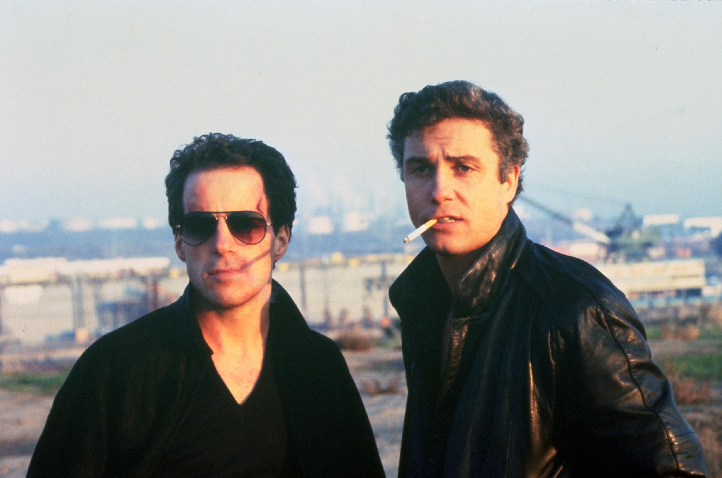 Two individuals stand outdoors against an industrial or urban backdrop; one wears a dark shirt and the other a dark jacket