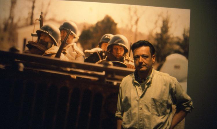 A large photo image of a group of people, some in helmets and uniforms, stand behind a barricade while a casually dressed individual stands in the foreground