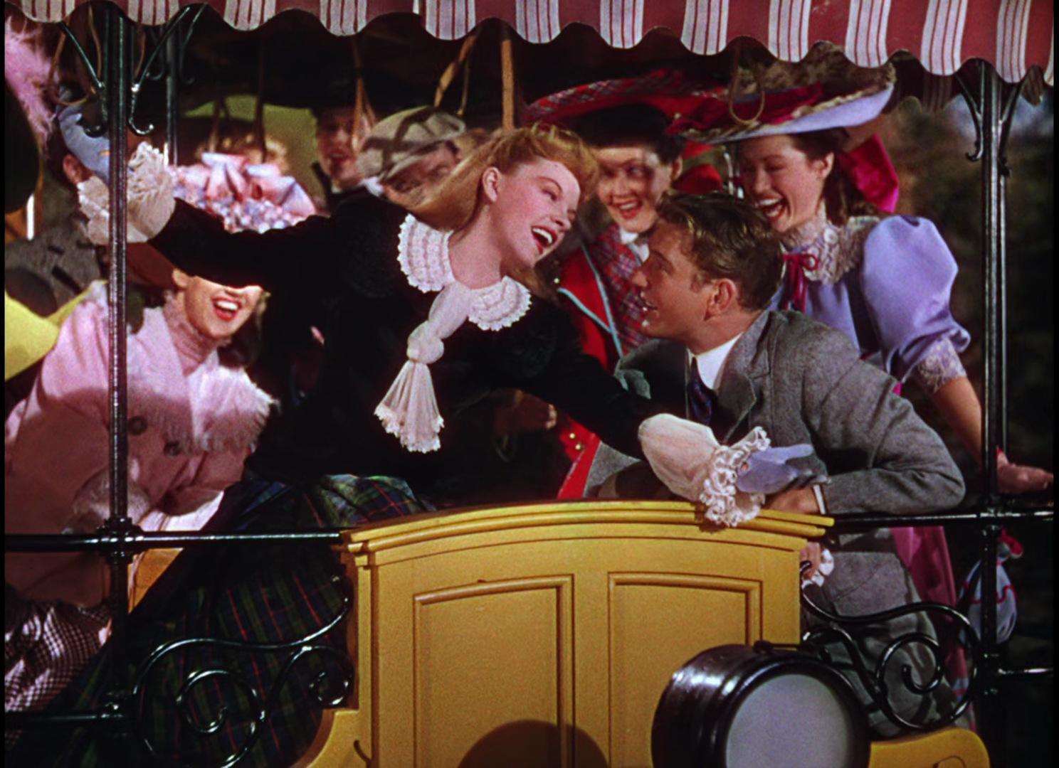 A group of people in late 19th or early 20th century costumes ride on an open carriage with a yellow front panel and decorative ironwork