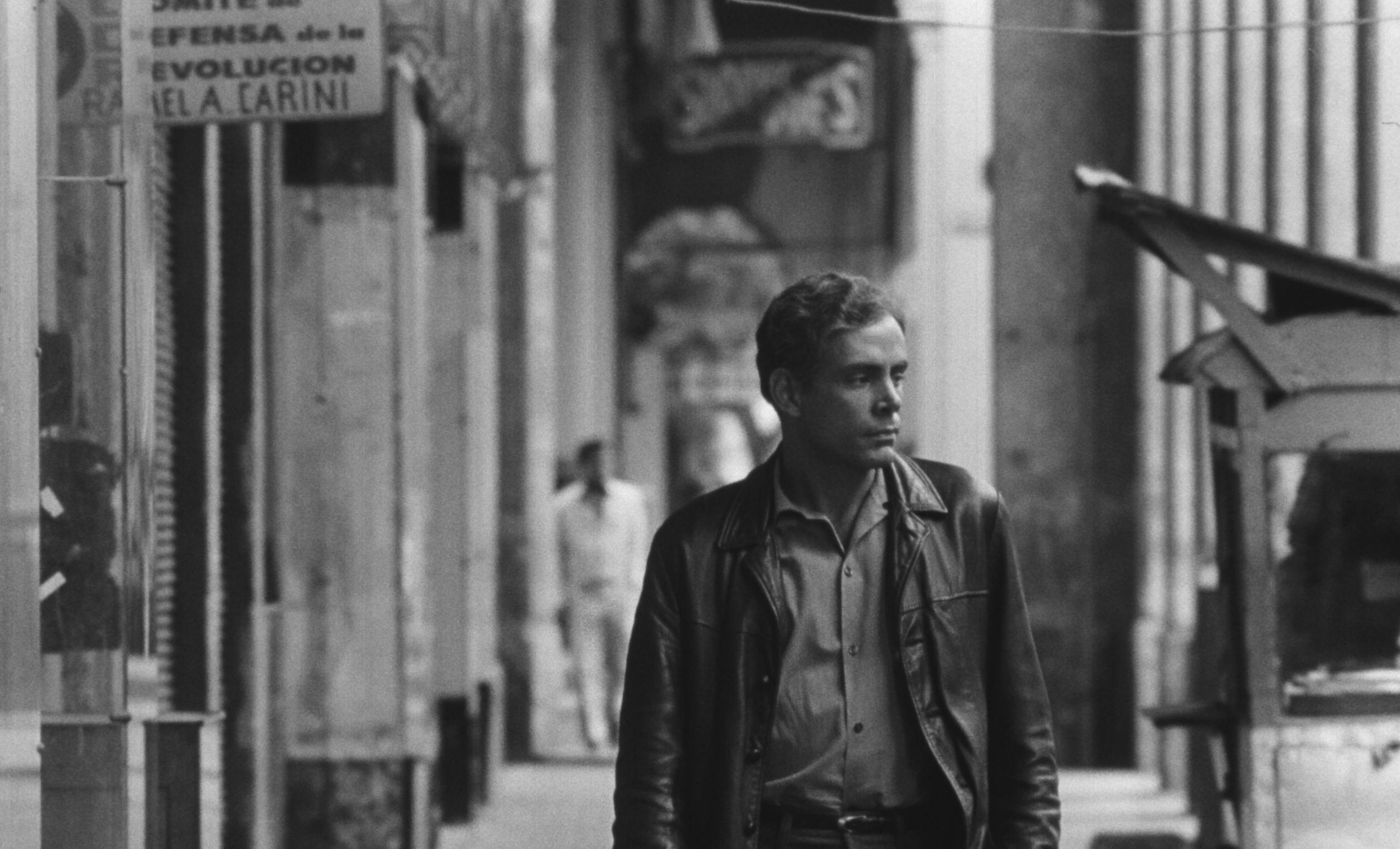 The image is black and white. A man in a leather jacket walks down an urban street with buildings, signs, and other pedestrians in the background.