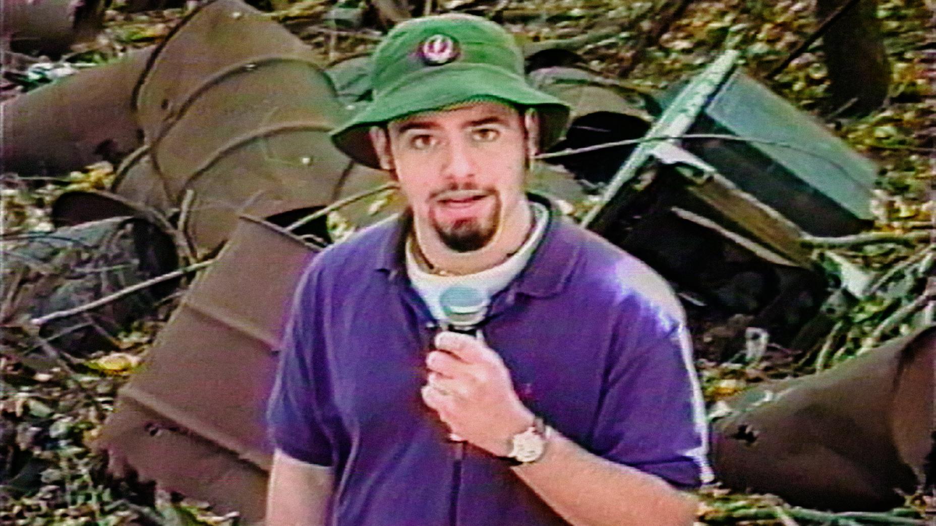 A person in a green hat and purple shirt holds a microphone in front of rusted metal barrels and debris.