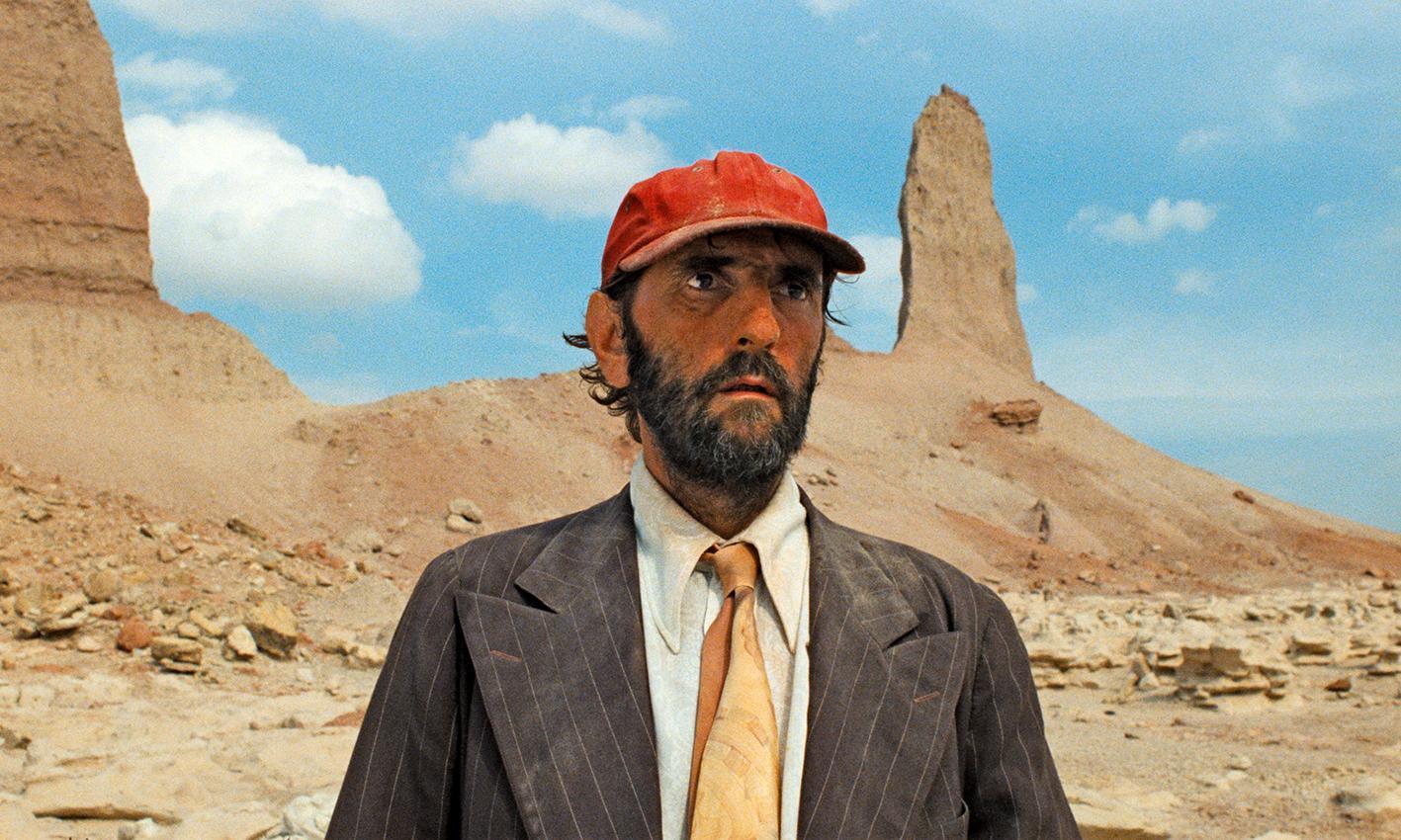 A person in a red cap, suit jacket, and orange tie stands in a desert landscape with rock formations in the background.