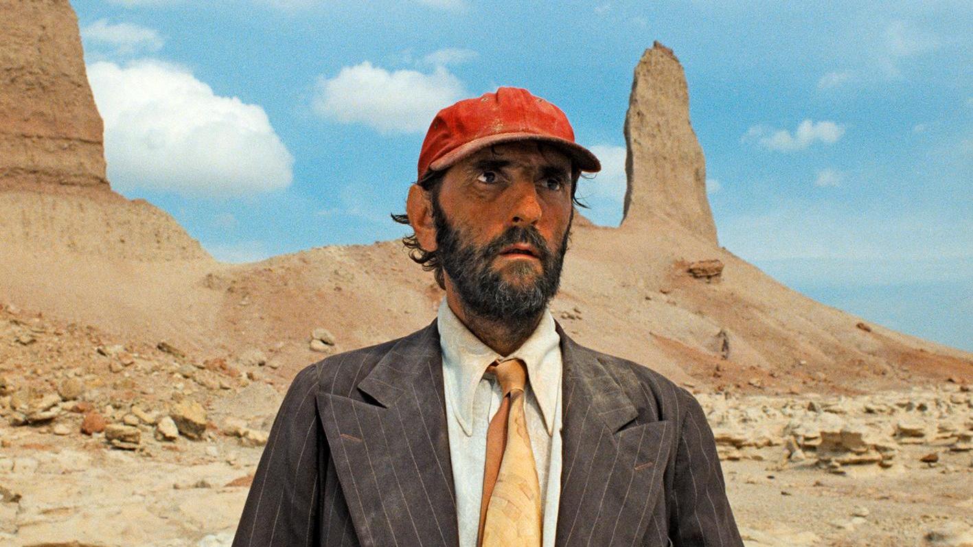 A person in a red cap, suit jacket, and orange tie stands in a desert landscape with rock formations in the background.