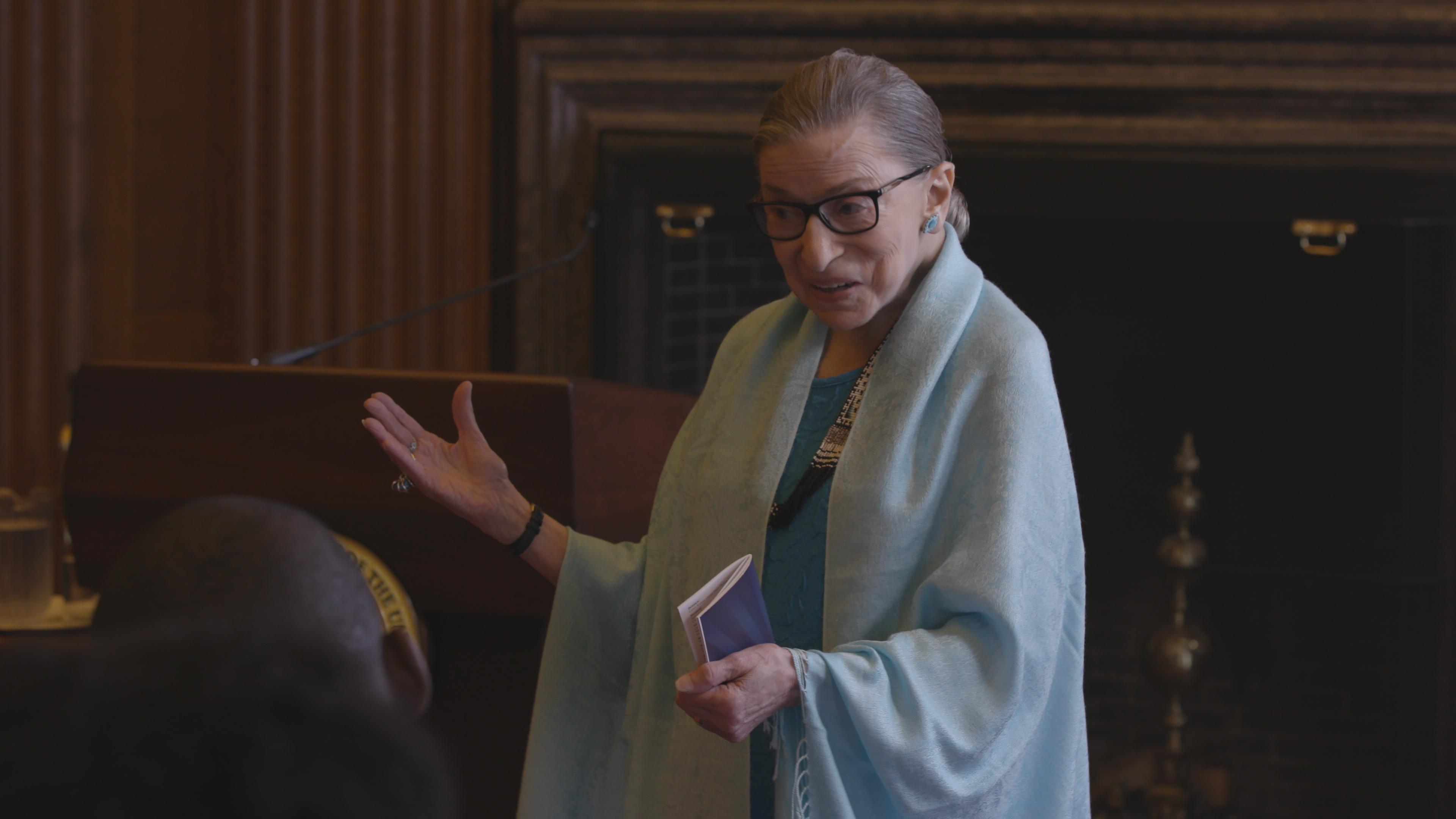 A person stands at a podium holding a small book and gesturing. The background features wooden paneling and a decorative fireplace