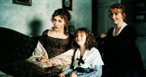 Three people in period clothing sit on a couch in a vintage setting with framed pictures and a door in the background