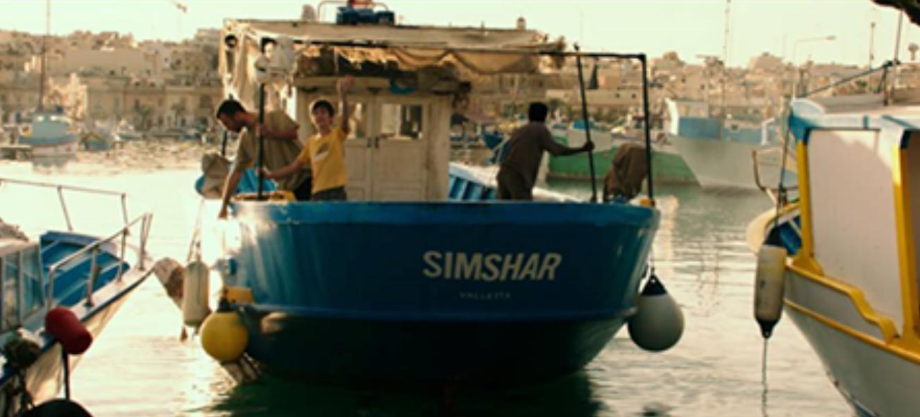 A blue fishing boat named 'SIMSHAR' is docked at a busy harbor with three people aboard; other boats and buildings are visible in the background