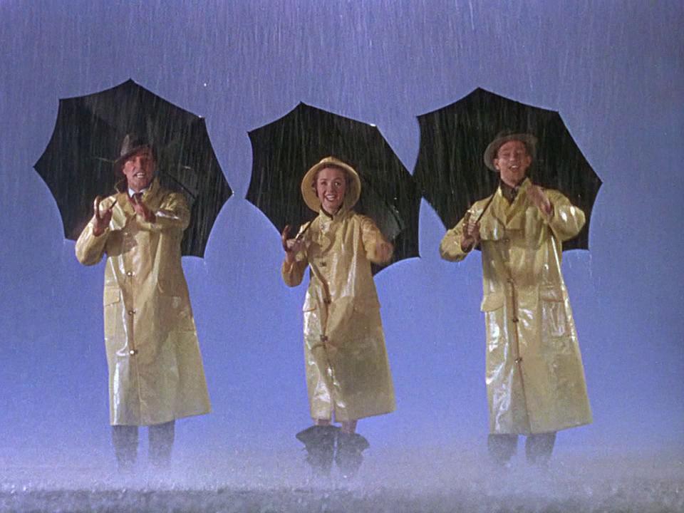 Three individuals in yellow raincoats stand in the rain holding black umbrellas; raindrops fall against a blue sky