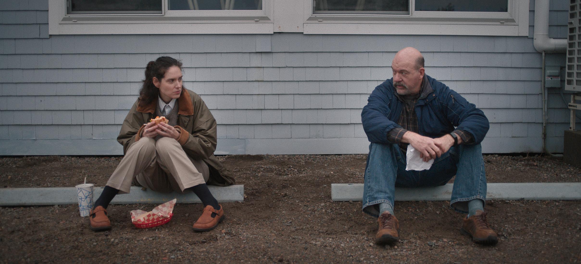 Two people sit on the ground outside a building; one is eating a sandwich while the other holds a napkin