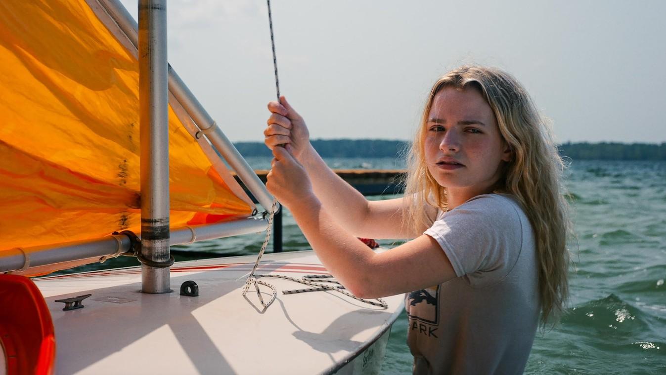 A young woman with long blonde hair sails a small sailboat with an orange sail on a lake.