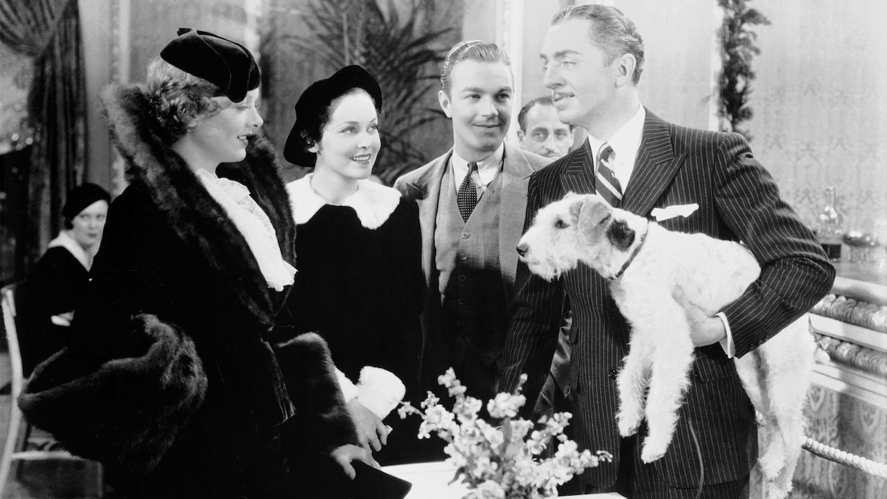 Black and white photo of four people in formal attire; the man on the right holds a small dog