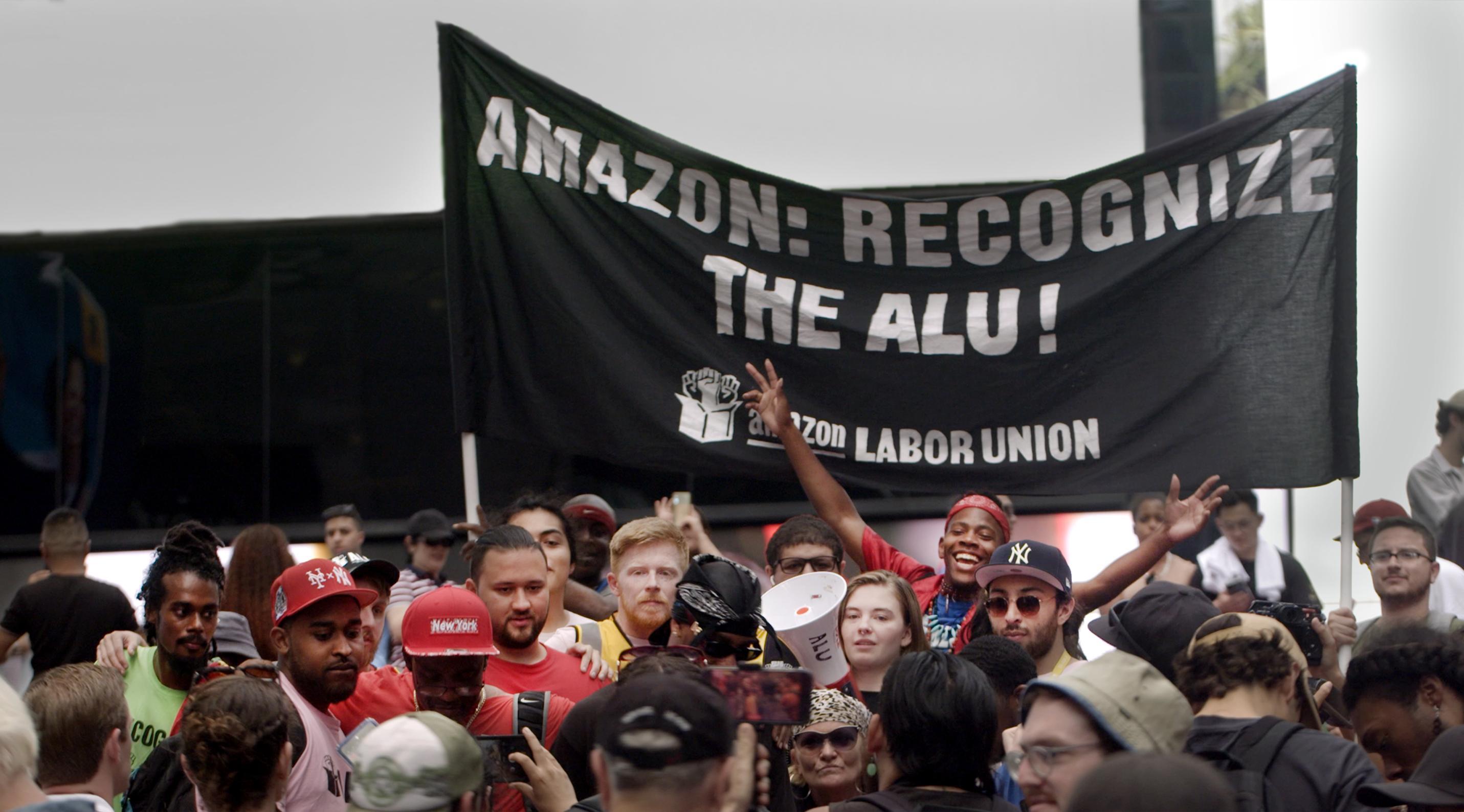 Group of people holding a large black banner that reads 'AMAZON: RECOGNIZE THE ALU!' with 'AMAZON LABOR UNION' and raised fists printed below