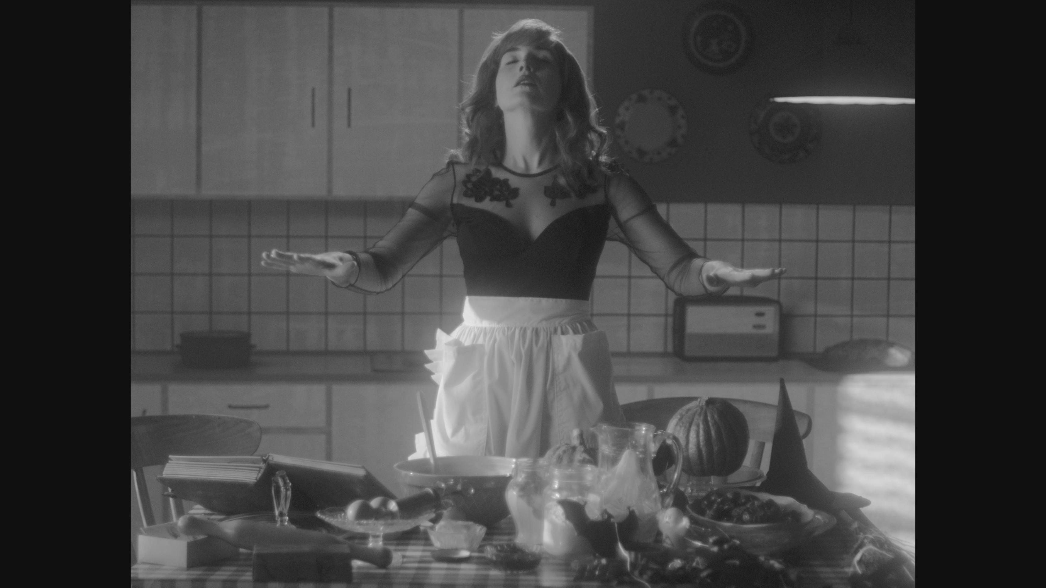 Person with arms outstretched stands in a kitchen wearing a black top with sheer sleeves and a white apron. The kitchen features tiled walls, cabinets, appliances, and a table with food items like bread, fruit, and baked goods