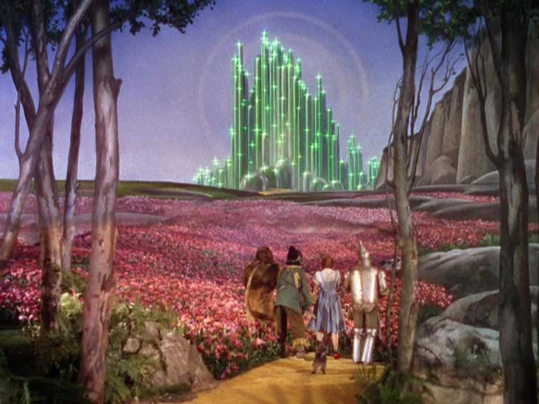 Scene from 'The Wizard of Oz' with Dorothy, the Scarecrow, Tin Man, and Cowardly Lion walking along the yellow brick road through red flowers toward the glowing green Emerald City