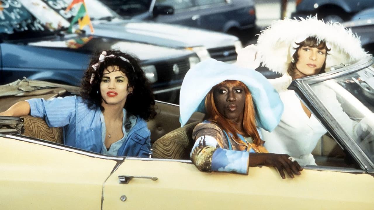 Three individuals sit in a yellow convertible; one wears a blue shirt, another a large blue hat and colorful clothing, and the third a white outfit with a white hat