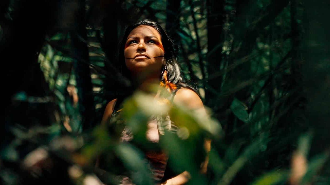Person standing in a dense forest with lush green foliage; their face is partially covered by leaves and branches