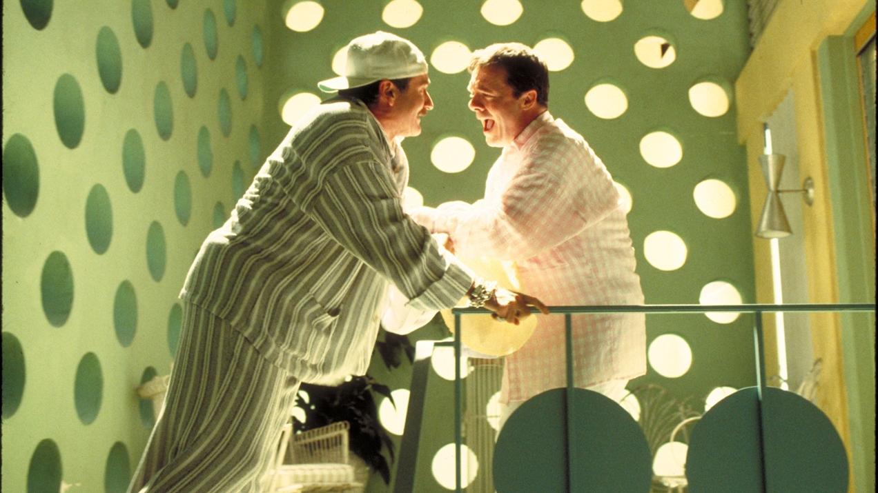 Two men wearing pastel striped pajamas laugh together in a room with polka dot walls.