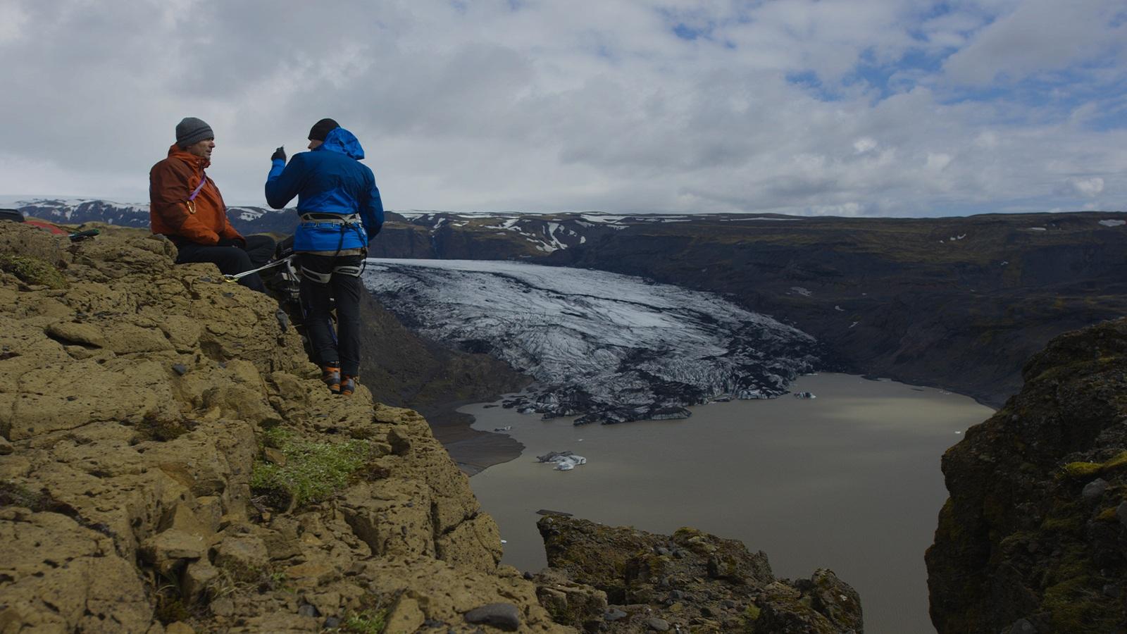 Two people in ski jackets perched on a mountain cliff high above melting glaciers and a lake.