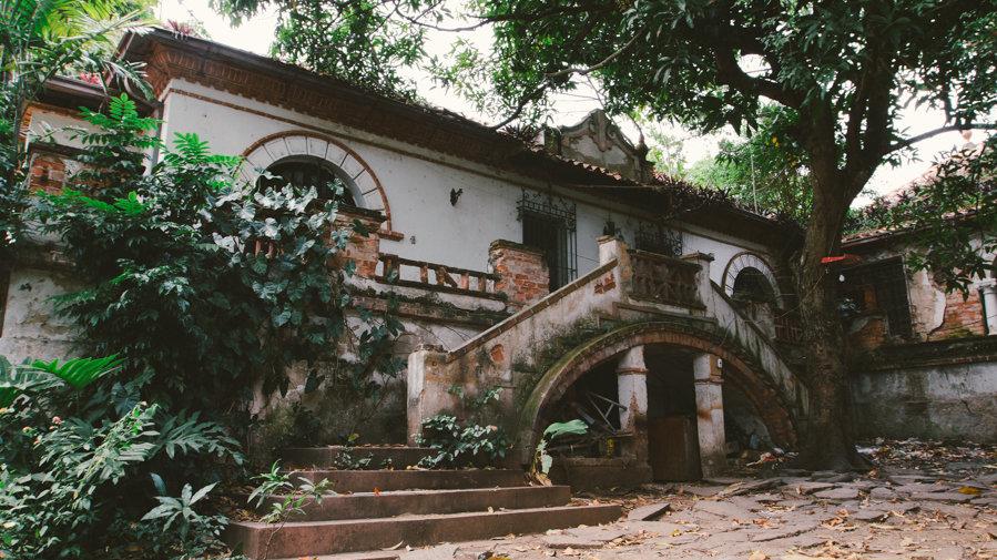 A dramatic photo of an seemingly abandoned two-story Spanish-style manor being overtaken by vines and trees.