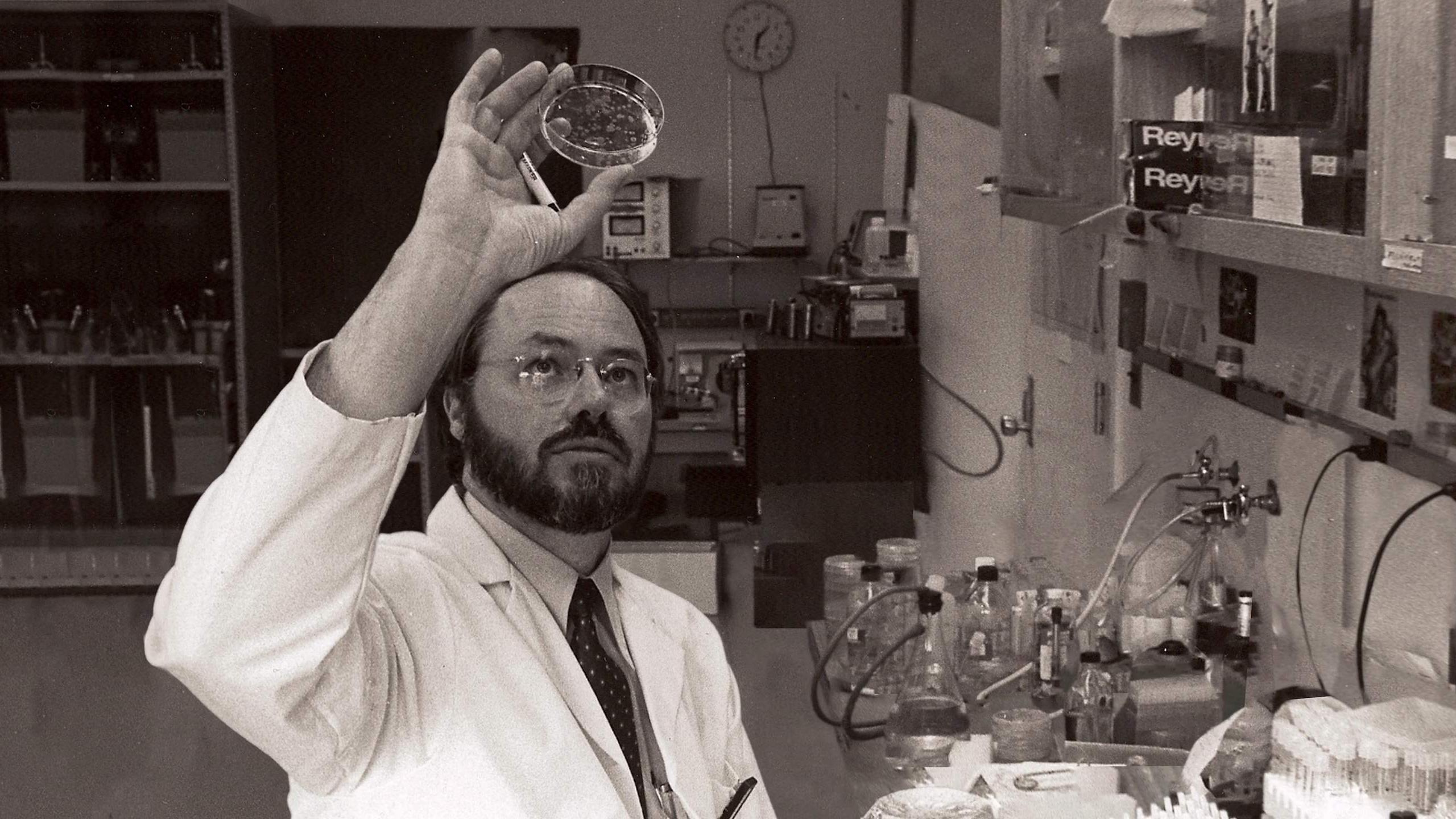 A black and white still image of a man in a white lab coat holding a petri dish up above his face to examine it.