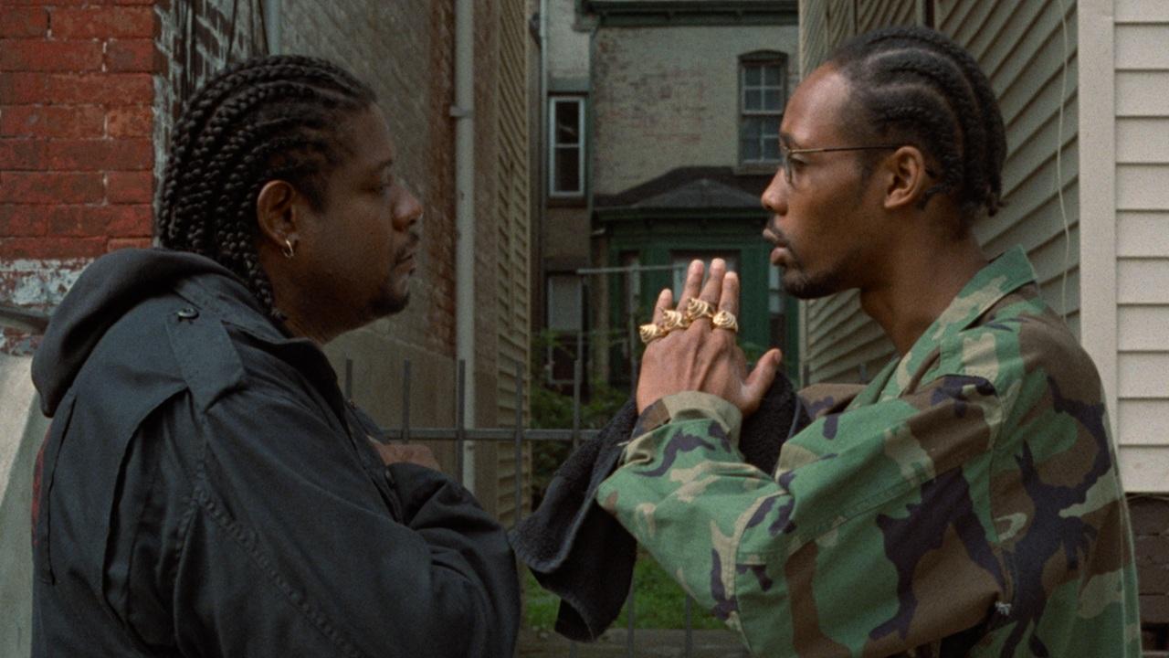 A man wearing a black jacket stands facing a man with his hands in a prayer gesture wearing a camo jacket and lots of gold rings.