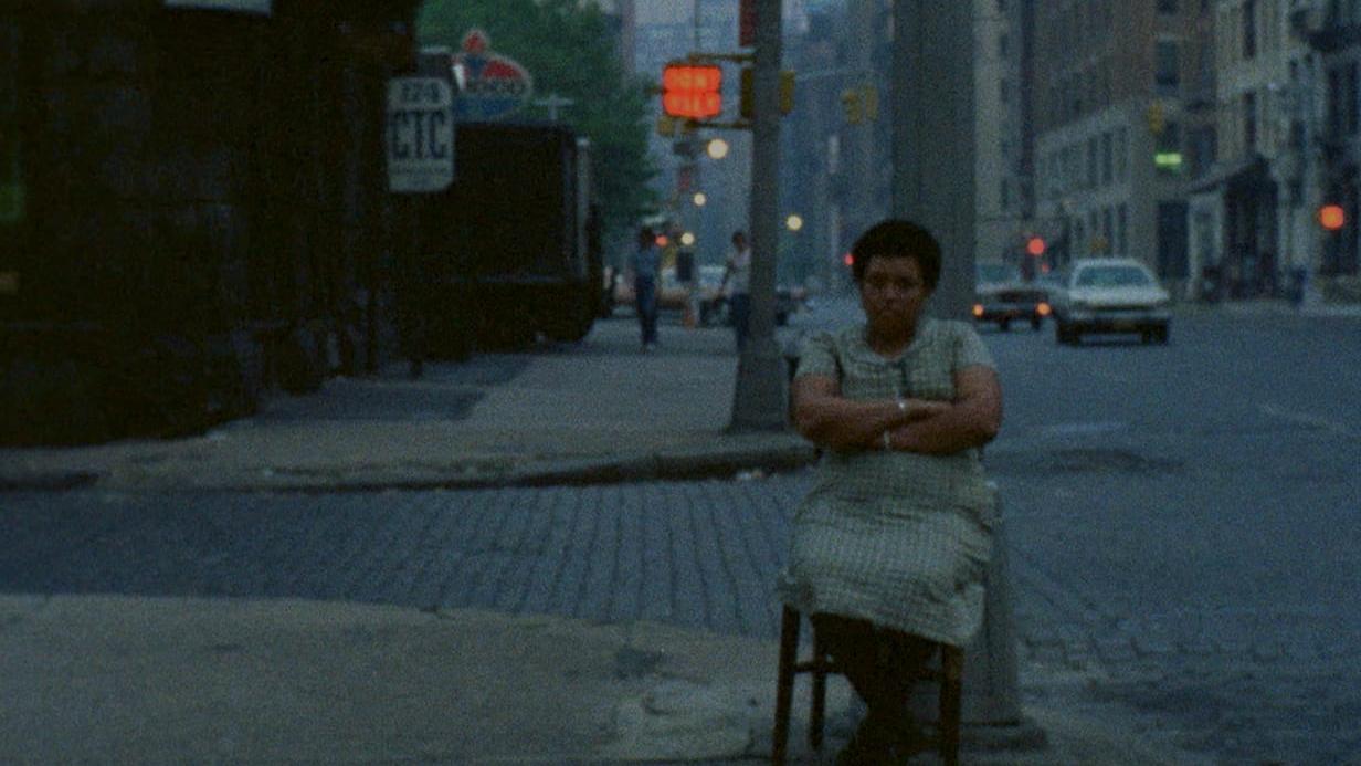 A woman sitting with her arms crossed in a wooden chair at a city intersection. There is a "Don't Walk" sign illuminated above her.