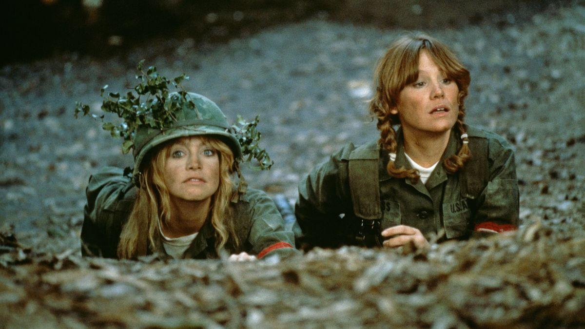 Two women lying on the ground wearing army uniforms. One has pigtail braids and one is wearing an army hat decorated with flowers.