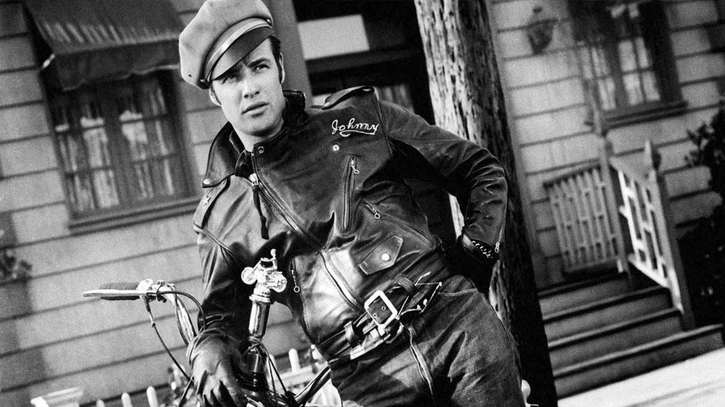 A man with a leather jacket and military cap leaning casually against a motorcycle.