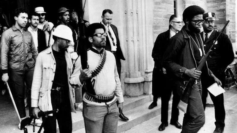 A group of young men exiting a building carrying rifles. The man at center is wearing bullets across his chest and around his waist.