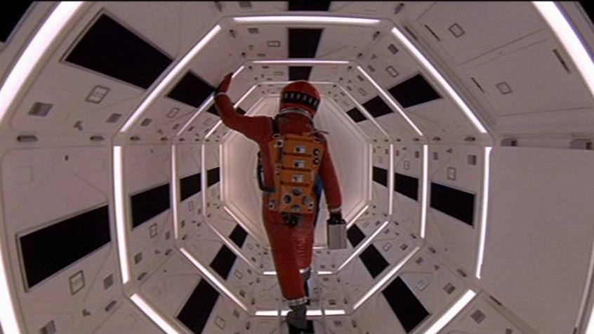 A person in a red space suit walks through a brightly lit, hexagonal corridor with futuristic design.