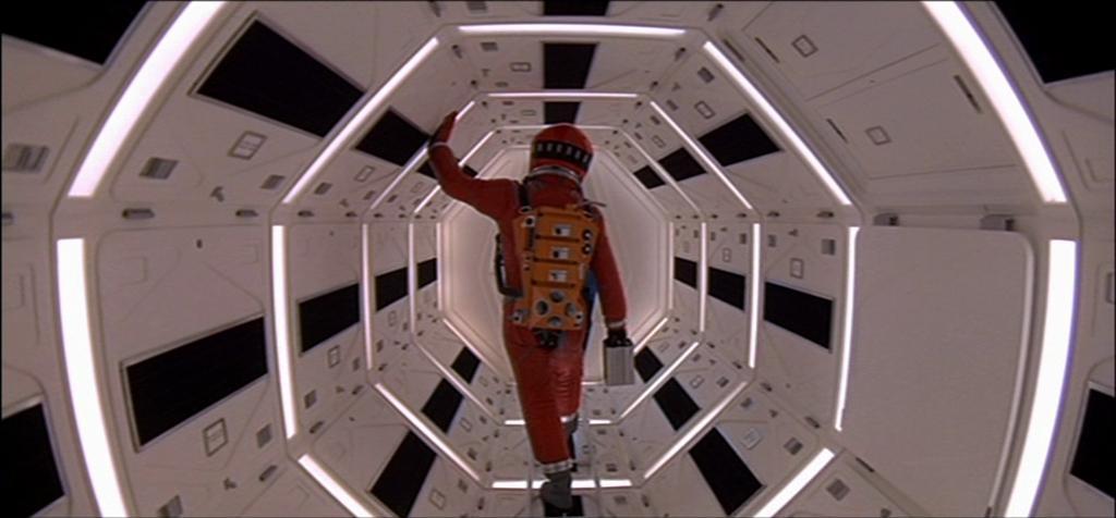 A person in a red space suit walks through a brightly lit, hexagonal corridor with futuristic design.
