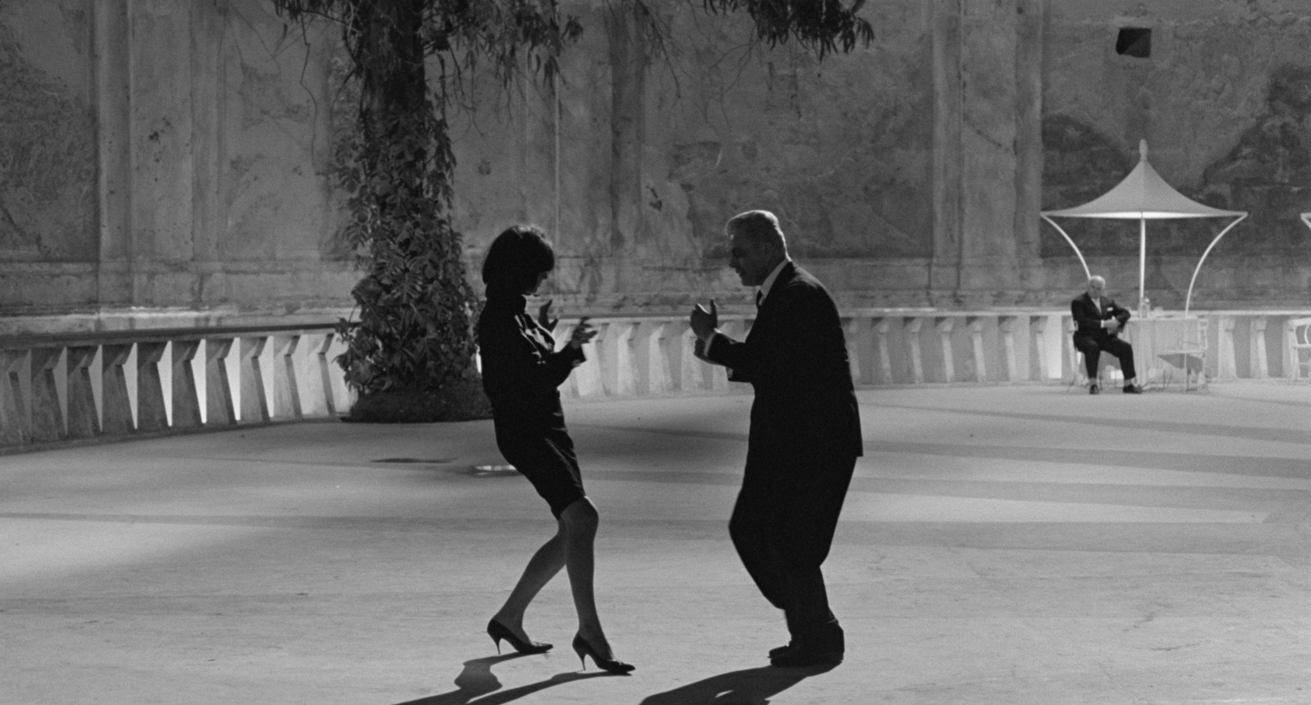 Two people dance energetically in an open courtyard, one in a dress and heels, the other in a suit. A third person sits at a table under a canopy, watching the scene unfold.
