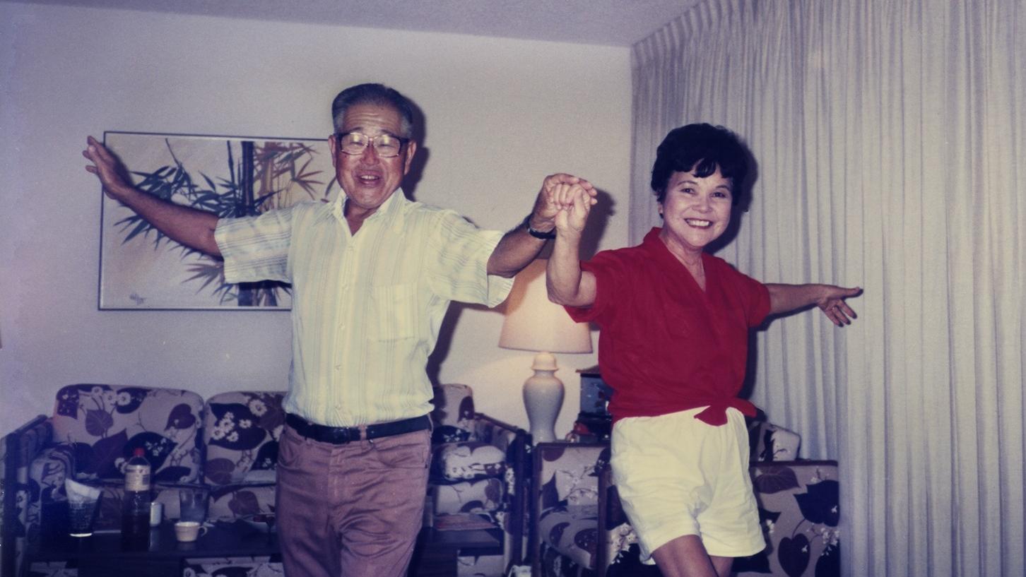 A vintage photograph of man wearing glasses dancing joyously in a living room with a woman in a red shirt. They are holding hands and their arms are up in the air.