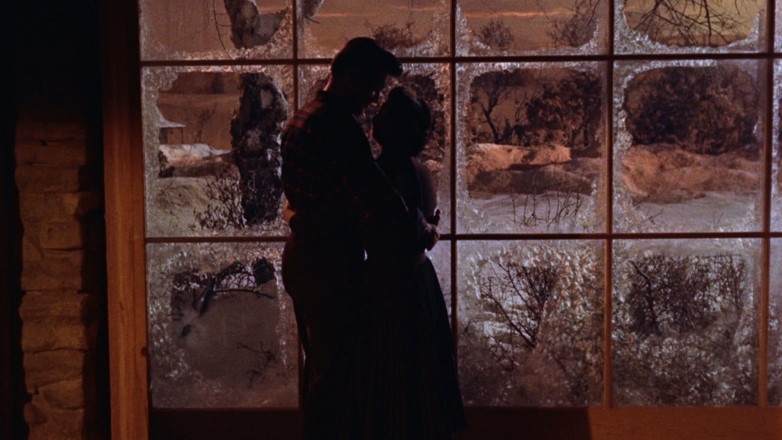 Two people stand closely together inside a building, silhouetted against a frosted window with a snowy landscape outside. Warm light filters through the panes, creating a contrast between the cold outdoors and the intimate indoor setting.