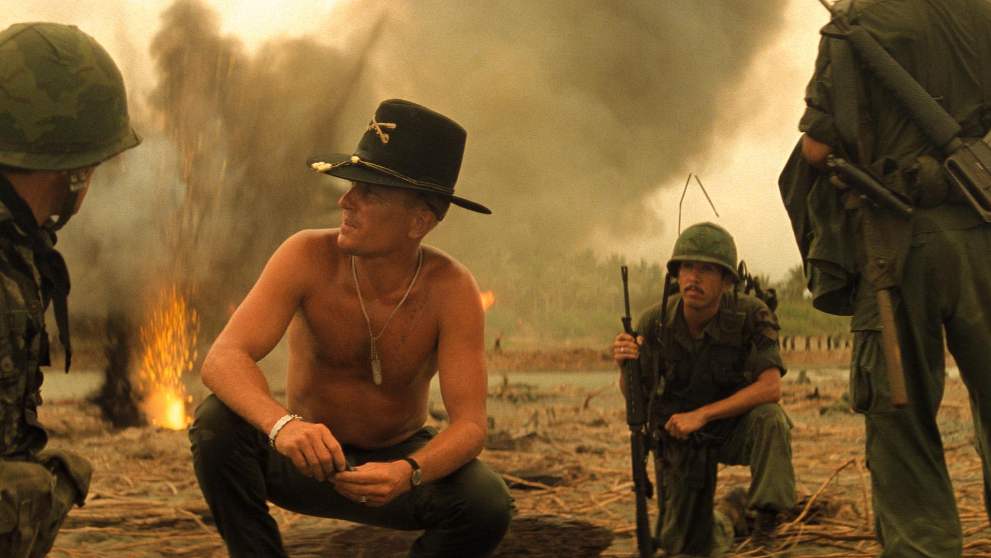 Soldiers gather in a smoky war zone with fire in the background, suggesting recent combat activity. A shirtless man in a cavalry hat speaks to another soldier, while others stand nearby in military gear.