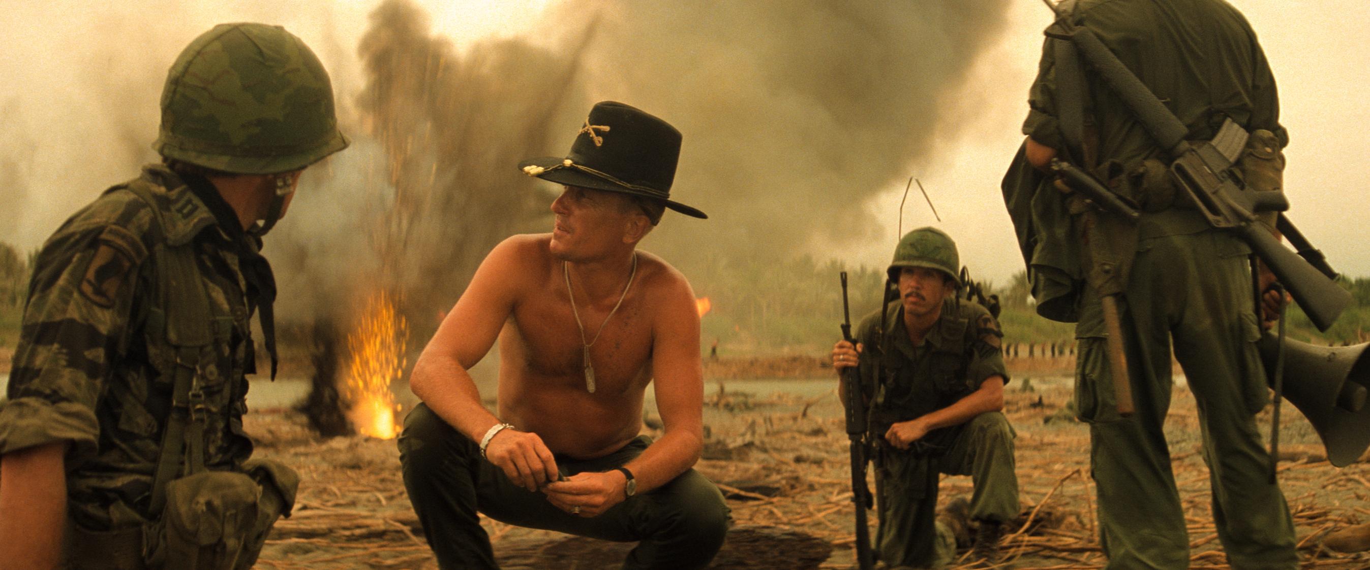 Soldiers gather in a smoky war zone with fire in the background, suggesting recent combat activity. A shirtless man in a cavalry hat speaks to another soldier, while others stand nearby in military gear.