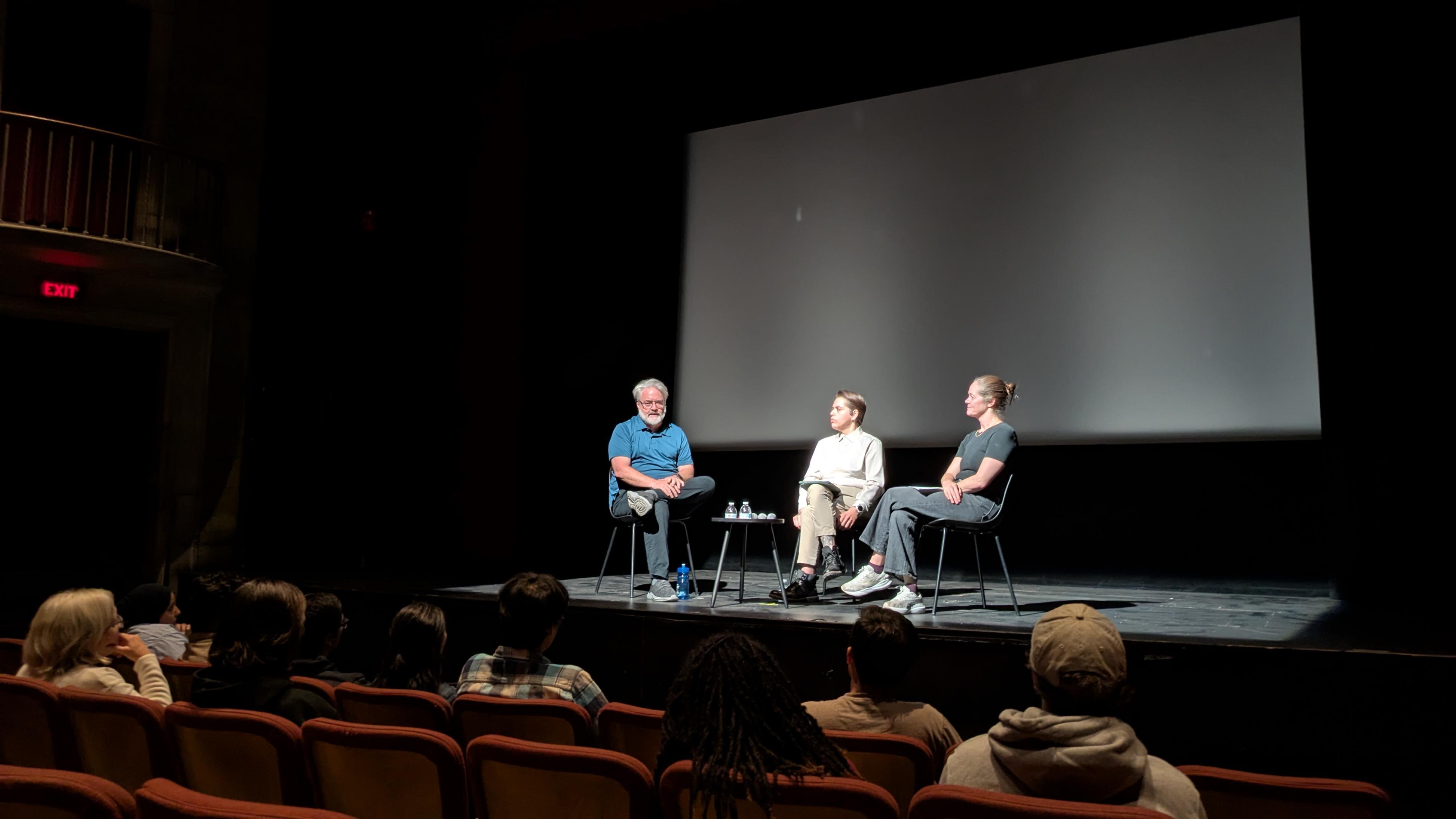 Three people sit on stage in a theater, engaged in a panel discussion or interview.