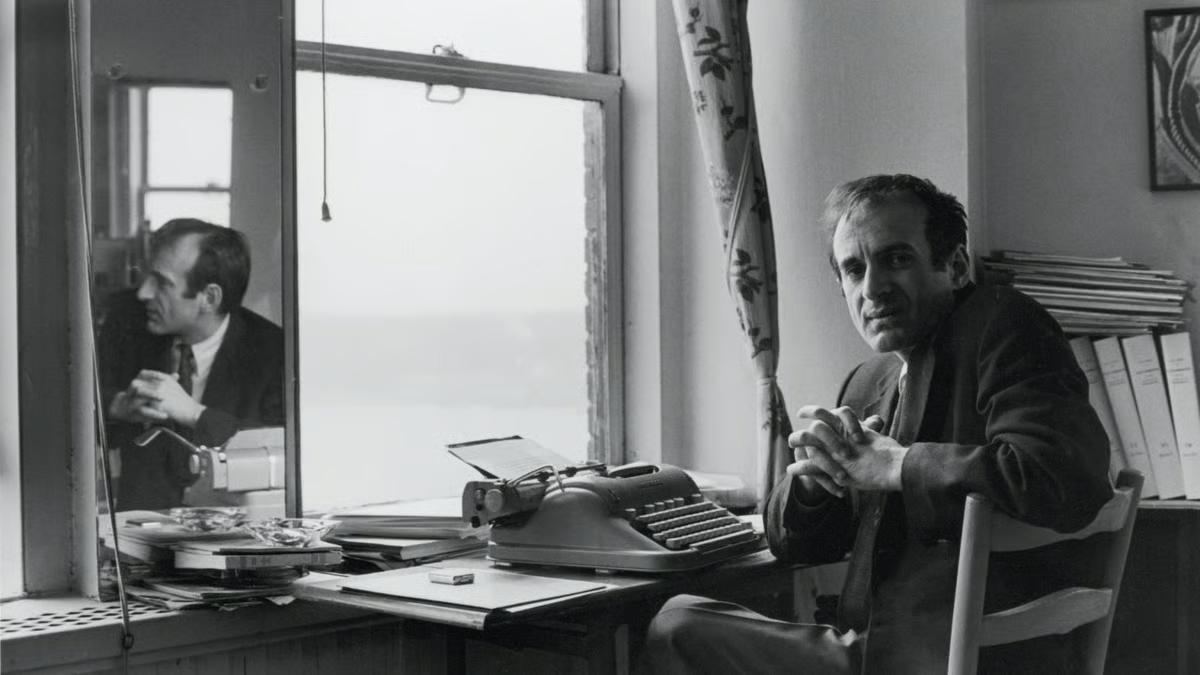  A man in a suit sits at a desk with a typewriter, papers, and books. Behind him, a window with floral curtains reveals a body of water. A mirror reflects his profile and part of the room, adding depth to the vintage office setting.