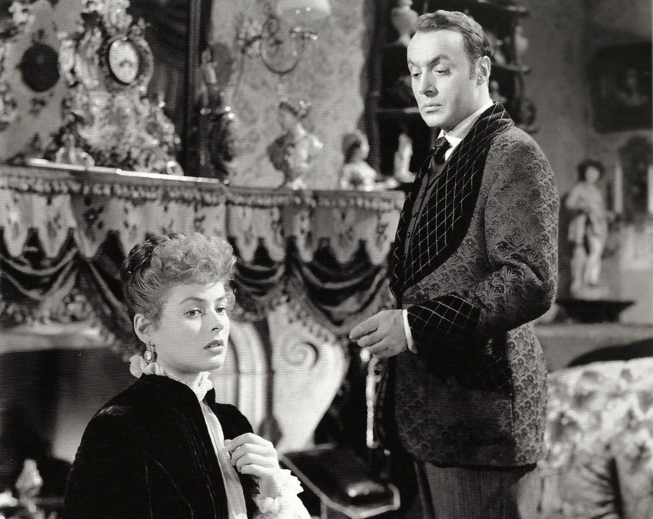 A woman in a high-collared blouse and earrings stands beside a man in a patterned velvet jacket and cravat. They are in a richly decorated Victorian room with statues, a large clock, and heavy drapery.