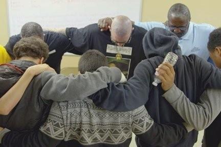 Eight people stand in a circle indoors with arms around each other’s shoulders, sharing a moment of unity. One person holds a microphone, and the background includes a whiteboard and beige walls, suggesting a classroom or community space.