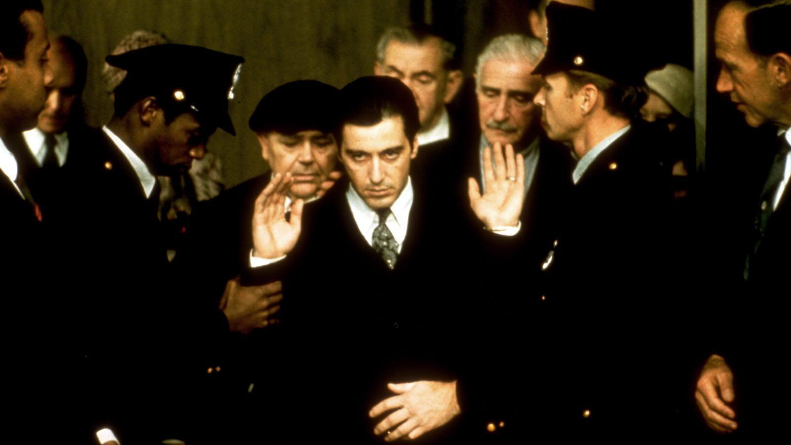 A group of people dressed in formal attire and uniforms are gathered closely together, with some raising their hands as if in greeting or acknowledgment. The scene appears to be part of an official or ceremonial event.