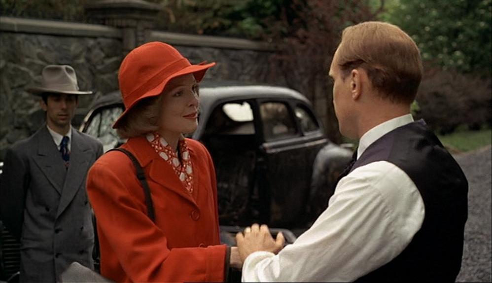 Two people are standing outdoors near a vintage car, with one person wearing a bright red coat and hat while holding hands with another person in a dark vest and white shirt.