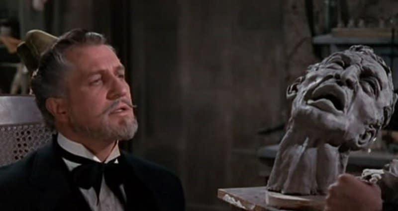 A man in a black suit and bow tie sits beside a dramatic sculpted bust with an anguished expression. The indoor setting includes shelves and tools, suggesting an artist’s studio or workspace.