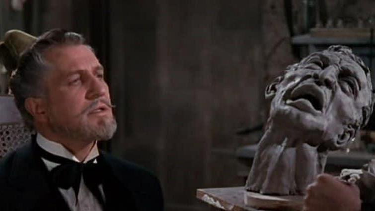 A man in a black suit and bow tie sits beside a dramatic sculpted bust with an anguished expression. The indoor setting includes shelves and tools, suggesting an artist’s studio or workspace.