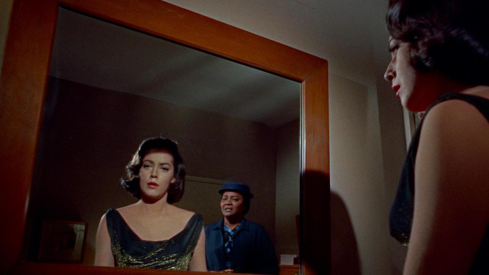 A woman in a black dress with a shimmering neckline looks into a mirror, revealing her reflection and a second person in a blue outfit and hat standing behind her. 