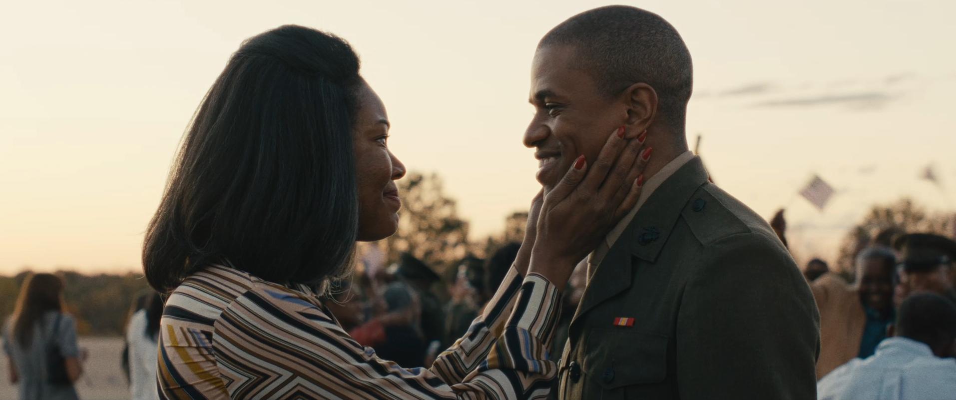 A person wearing a striped shirt gently touches the face of another person dressed in a military uniform during an outdoor gathering. People and small flags are visible in the background under a clear sky.