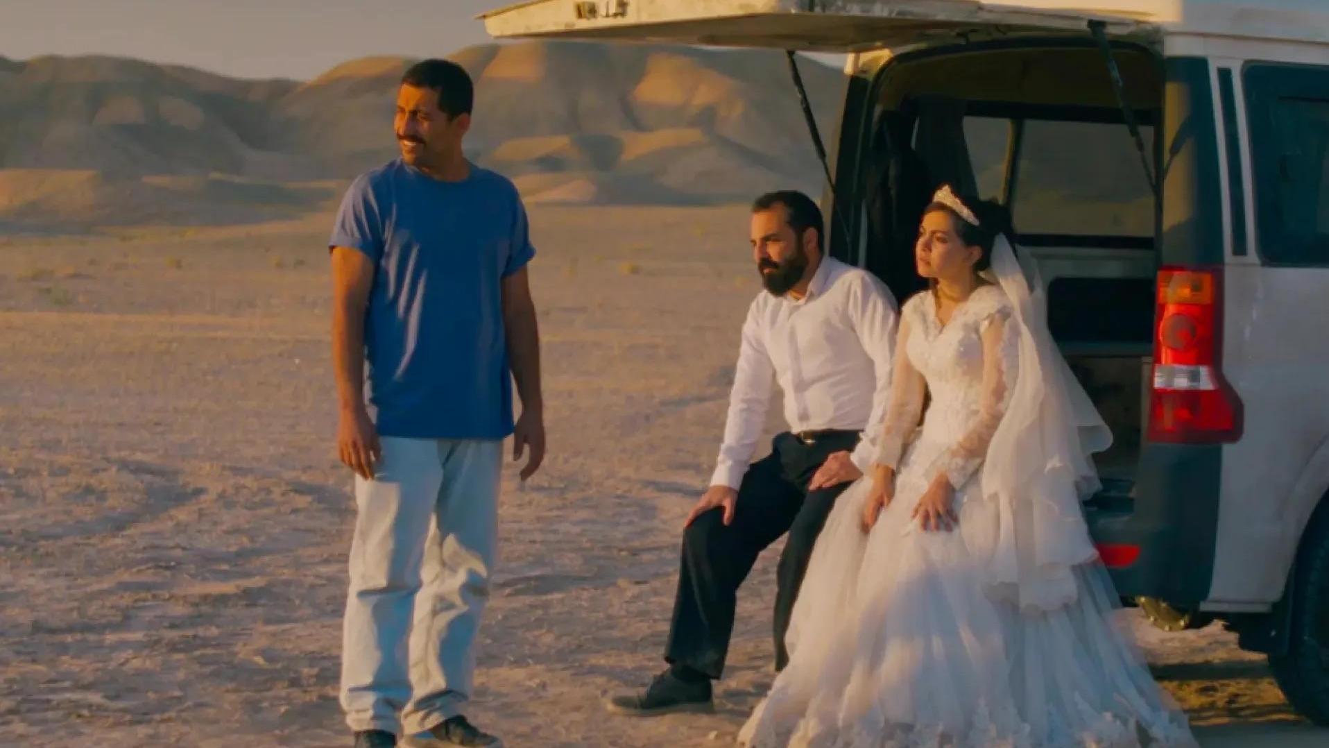 A man in a white shirt and dark pants sits beside a woman in a white wedding dress at the open back of a silver van. Another person stands nearby in a blue shirt and light pants. The desert-like landscape and mountains in the background contrast with the formal wedding attire.