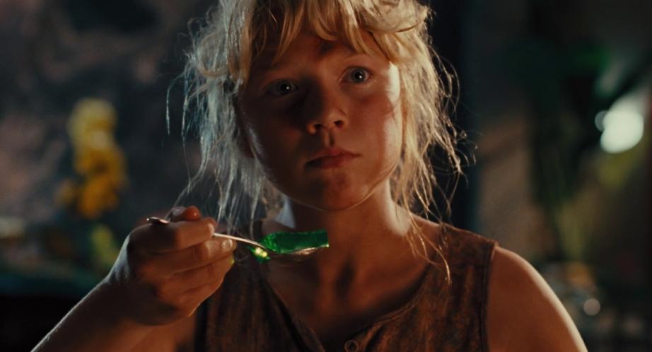 A young person with light hair holds a spoon with green gelatin, gazing intently off-camera. Dim lighting and blurred background elements, including a yellow flower and greenery, create a dramatic and contemplative atmosphere.