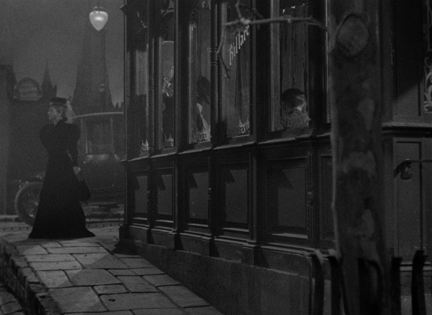 A woman in a long coat and hat walks along a cobblestone sidewalk at night past a vintage storefront with ornate windows. Inside, people are visible through the glass, and a sign partially reads “Billiard.” A streetlamp casts light on the scene, with a vi